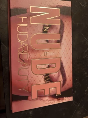 Huda Beauty New Nude ögonskuggspalett - Säljer en Huda Beauty New Nude ögonskuggspalett med 18 olika nyanser i matta, skimriga och glittriga toner. Färgerna går i nude, rosa, brunt, guld och lila. Paletten har en snygg rosa förpackning med spegel och magnetstängning.