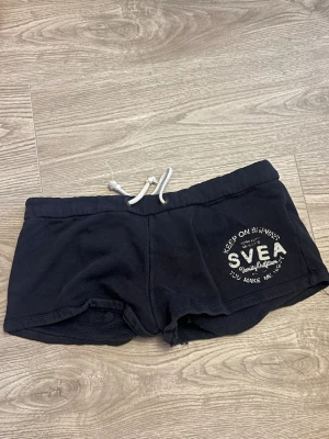 Svarta shorts med snörning från SVEA - Svarta mjuka shorts från SVEA med vit snörning i midjan och tryckt logga på ena benet. Shortsen har en bakficka och är perfekta för sommarens alla äventyr. Sköna och avslappnade med normal passform.
