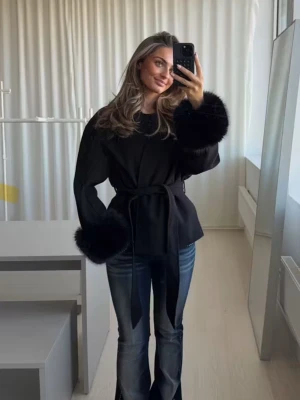 Svart kappa med fuskpälsdetaljer - Snygg svart kappa med markerad midja och knytskärp. Ärmsluten har lyxiga fuskpälsdetaljer som ger en trendig look. Perfekt för dig som vill sticka ut och hålla stilen under kyliga dagar. Passar till jeans eller klänning.