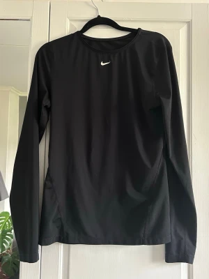 Svart långärmad träningströja Nike - Svart långärmad träningströja från Nike med rund hals och diskret vitt logotryck på bröstet. Tillverkad i ett lätt och stretchigt funktionsmaterial med ventilerande meshpaneler i sidorna för extra andningsförmåga. Perfekt för löpning eller gym.