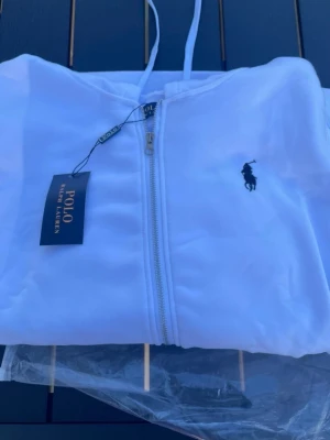 Vit hoodie från Polo Ralph Lauren - Snygg vit hoodie från Polo Ralph Lauren med dragkedja framtill och klassisk logga broderad på bröstet. Hoodien har huva med dragsko och är tillverkad i mjukt material som känns skönt mot huden. Perfekt för en clean och stilren look.