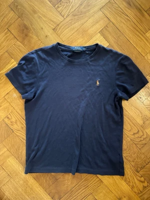 Mörkblå t-shirt från Polo Ralph Lauren - Klassisk mörkblå t-shirt från Polo Ralph Lauren med korta ärmar och rund hals. T-shirten har den ikoniska broderade loggan på bröstet och är gjord i mjuk bomull för en skön känsla. Perfekt till jeans eller shorts.
