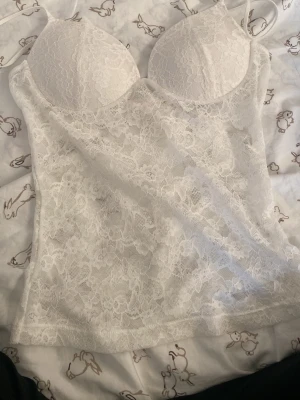 Vit spetsbody med vadderade kupor - Säljer en vit linne genomskinlig spets med vadderade kupor och smala axelband.  har ett blommigt spetsmönster och ger en feminin och elegant look. Perfekt för dig som gillar detaljer och mjuka material.