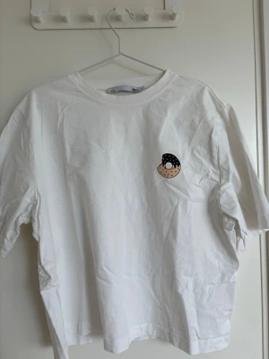 Vit t-shirt med tryck från 157 - Vit t-shirt från 157 med ett litet tryck av en tecknad figur på bröstet. Klassisk rund hals och korta ärmar. Perfekt för dig som gillar enkel stil med en rolig detalj. Tillverkad i mjuk bomull för skön känsla hela dagen.