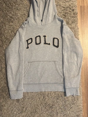 Grå hoodie med POLO-tryck - Ljusgrå hoodie med stor huva och magficka. Framtill står det POLO i stora bokstäver med svart och beige brodyr. Tillverkad i mjukt sweatshirtmaterial, perfekt för chill dagar. Klassisk passform och ribbade muddar vid ärmar och nederkant. Har ett litet hål vid bröstet där av priset men syns knappast, annars jätte fin. 