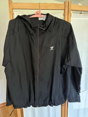 Svart vindjacka med huva från Adidas - Snygg svart vindjacka från Adidas med dragkedja framtill, justerbar huva och dragsko i nederkant. Jackan har Adidas-logga på bröstet och är tillverkad i lätt, vindtätt material. Perfekt för blåsiga dagar och enkel att matcha med olika outfits.
