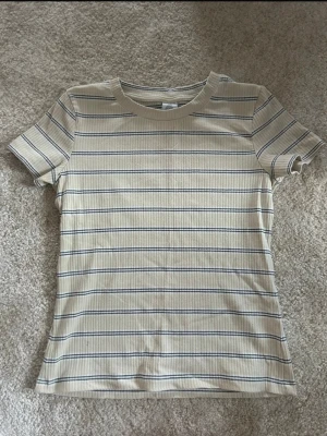 Randig tröja  - Säljer en gul t-shirt med tunna blå horisontella ränder. T-shirten har rund halsringning och korta ärmar. Passformen är normal och materialet känns mjukt, troligtvis bomull. Perfekt till jeans eller kjol.