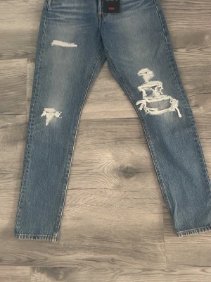 Blå slitna raka jeans - Säljer ett par blå jeans med raka ben och flera slitningar på framsidan. Jeansen har en klassisk femficksmodell och är tillverkade i denim. Perfekta för en avslappnad och trendig look. 27 storlek och längd 30