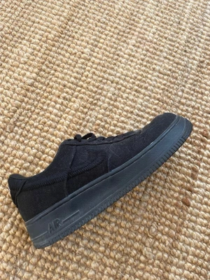 Svarta Stüssy x Nike Air Force 1 - Säljer ett par svarta Stüssy x Nike Air Force 1 sneakers i tygmaterial med matchande svarta skosnören och sula. Snygg broderad Swoosh på sidan och Stüssy-logga på plösen. Perfekta för dig som gillar streetwear och klassisk design.