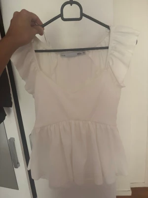 Vit volangblus med kort ärm - Söt vit blus med volangdetaljer och korta ärmar. Blusen har en luftig passform och är tillverkad i ett lätt material som passar perfekt till sommaren. Den har en feminin look med volanger vid axlarna och en något utställd nederdel.