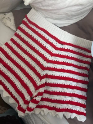 Randiga stickade shorts i rött och vitt - Säljer ett par stickade shorts med röda och vita ränder. De har hög midja, ribbad linning och vågig kant nertill för en söt touch. Perfekta för sommarens chill dagar eller som mysplagg hemma.