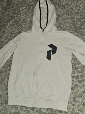 Vit PeakPerformance hoodie - Snygg vit PeakPerformance hoodie med dragkedja och svart grafiskt tryck på bröstet. Hoodien har huva med svart kant, två fickor framtill. Perfekt för en chill och avslappnad stil. Inga deffekter. Storlek S. OPS! Jag kan bara skicka med PostNord.