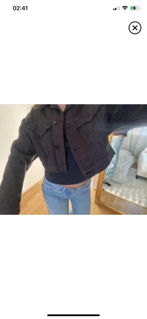Svart croppad jeansjacka - Säljer en svart croppad jeansjacka med klassisk krage och knappar framtill. Jackan har två bröstfickor med lock och långa ärmar. Perfekt att slänga över en t-shirt eller hoodie för en chill och snygg look.