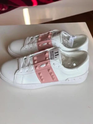Vita sneakers med rosa nitar - Snygga vita sneakers från Valentino med bred rosa rem över vristen dekorerad med pyramidformade nitar. Skorna har vita snören, rund tå och detaljerad sula. Tillverkade i slätt läder för en clean och modern look.