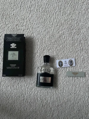 Creed Aventus parfym 100 ml - Lyxig parfym från Creed, modellen Aventus, i en elegant genomskinlig glasflaska med svart och silverdetaljer. Flaskan rymmer 100 ml och kommer med originalkartong och tillhörande kort. Doften är ikonisk och exklusiv.