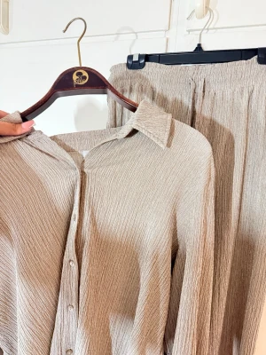Beige ribbat set med skjorta och byxa - Säljer ett snyggt set i beige med ribbad struktur. Setet består av en långärmad skjorta med krage och knappar framtill samt matchande vida byxor med resår i midjan. Så skönt set och stretchigt, älskar färgen på dom men använder tyvärr inte så mycket färg så det ligger mest i garderoben. Skjortan är helt oanvänd men byxorna använda vid 2/3 tillfällen kanske. Pris går att diskutera 💕