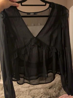 Svart  blus med volanger - Snygg svart transparent blus från Bik Bok i storlek XS. Blusen har långa ärmar, djup v-ringning och volangdetaljer framtill. Använd vid enbart ett tillfälle 