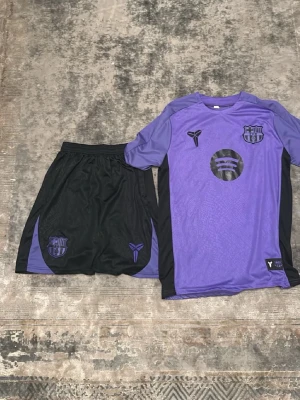 Lila och svart FC Barcelona träningsset - Säljer ett snyggt träningsset från FC Barcelona i lila och svart. Setet består av en kortärmad tröja med klubbmärke och sponsorlogga samt matchande shorts med elastisk midja och tryckta detaljer. Perfekt för fotbollsträning eller gymmet.