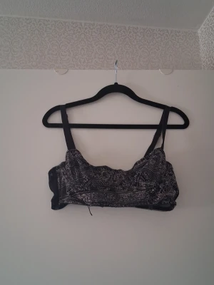 Svart bralette med mönster - Säljer en svart bralette med tunna axelband och ett detaljerat vitt paisleymönster på framsidan. Baksidan är enfärgad svart med hakknäppning. Mjuk och bekväm modell utan bygel, perfekt för en avslappnad stil.