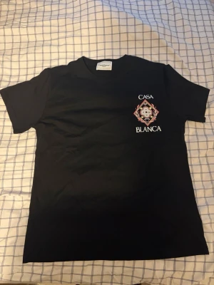  Casa Blanca T-Shirt - Snygg svart t-shirt från Casa Blanca med ett färgstarkt tryck och logga på bröstet. T-shirten har rund hals och korta ärmar. Materialet ser ut att vara mjukt bomullstyg, perfekt för en clean och stilren look.