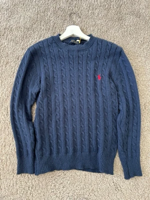 Mörkblå kabelstickad tröja Polo Ralph Lauren - Kabelstickad mörkblå tröja från Polo Ralph Lauren med rund halsringning och röd broderad logga på bröstet. Tröjan har långa ärmar och ribbade muddar vid ärmslut och nederkant. Perfekt för lager på lager och klassisk stil.
