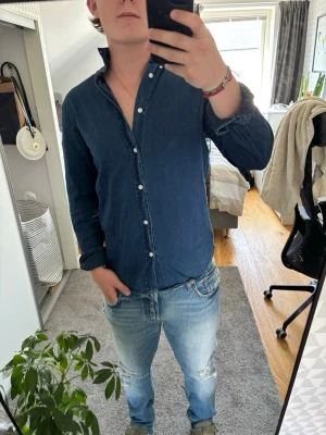 Mörkblå jeansskjorta med knappar - Säljer en klassisk mörkblå jeansskjorta med långa ärmar och knappar framtill. Skjortan har en avslappnad passform och är tillverkad i mjukt bomullsmaterial. Perfekt att styla lager på lager eller bära som den är.