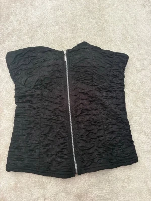 Svart tubtopp med dragkedja - Säljer en svart tubtopp med vågig struktur och dragkedja framtill. Toppen är axelbandslös och har en cool, croppad look som passar perfekt till jeans eller kjol. Materialet känns mjukt och följsamt mot huden.