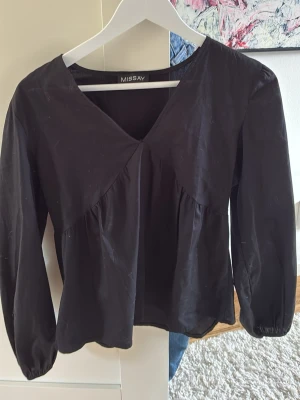 Svart blus med v-ringning från Missay - Stilren svart blus från Missay med v-ringad hals och lätt puffade långärmar. Blusen har en lös passform och fina sömdetaljer framtill som ger en snygg siluett. Perfekt att matcha med jeans eller kjol för en trendig look.