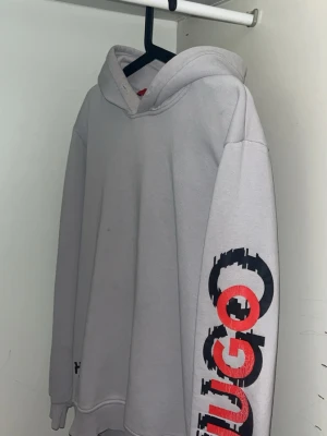 Ljusgrå hoodie med tryck från Hugo - Denna hoodie passar mer som M. Ljusgrå hoodie från Hugo med stor huva och ribbade muddar. Ärmen har ett stort svart och rött tryck med texten 'Hugo' i grafisk stil. Materialet är mjukt och bekvämt, perfekt för en avslappnad look. Liten logga även vid nederkanten. Liten håll och en fast märke på den 