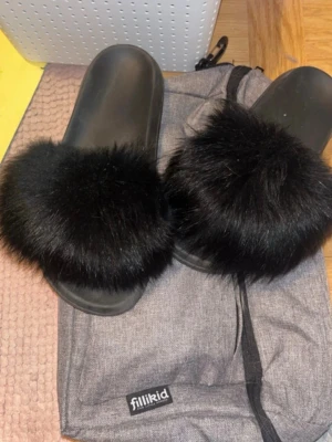 Svarta fluffiga tofflor - Säljer ett par svarta tofflor med bred rem täckt av mjukt, fluffigt fuskpäls. Sulan är i svart plast och designen är enkel men trendig. Perfekta för dig som vill ha något bekvämt och snyggt att glida runt i hemma.