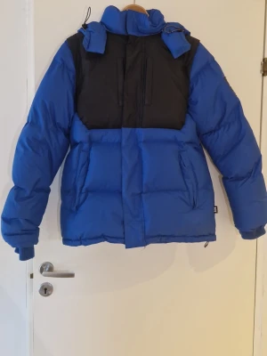 Blå och Svart Helly Hansen Collab med Sweet SKTBS - Säljer en blå pufferjacka med svarta partier över axlar och bröst. Jackan har en stor huva, dragkedja framtill och två bröstfickor med dragkedja. Puffig modell som håller värmen och har muddar vid ärmsluten. Perfekt för kalla vinterdagar. Jackan har även en innerficka och man ser lite användning på muddarna.