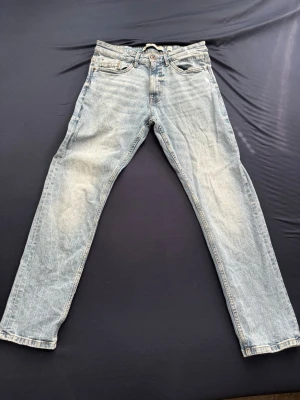 Ljusblå raka jeans - Säljer ett par ljusblå jeans med rak passform och klassisk femficksdesign. Jeansen har slitna detaljer och en vintage tvätt som ger en avslappnad look. Tillverkade i denim med normal midja och dragkedja fram.