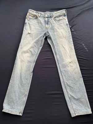 Calvin Klein jeans med slitningar - Ljusblå jeans med raka ben och diskreta slitningar på låren. Klassisk femficksmodell med orange sömmar och knappgylf. Jeansen har en avslappnad vintagekänsla och är tillverkade i denim.