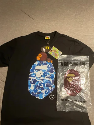 Svart BAPE t-shirt med blått camotryck - Säljer en svart t-shirt från A Bathing Ape med stort tryck av BAPE-apan och blått camomönster på framsidan. Klassisk rund hals och korta ärmar. Perfekt för dig som gillar streetwear och vill sticka ut med en ikonisk design.