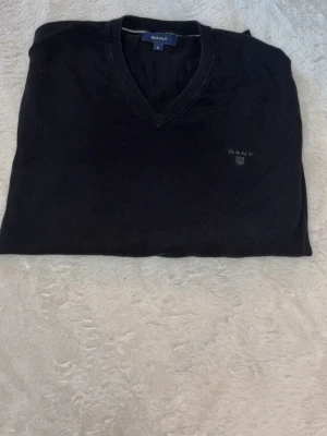 Svart V-ringad tröja från GANT - Klassisk svart V-ringad tröja från GANT i premium bomull. Diskret logga broderad på bröstet och ribbade muddar vid halsen. Perfekt för en clean och stilren look.