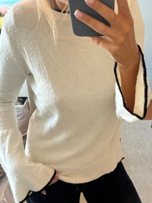 Vit tröja med svarta kanter - Säljer en stilren vit tröja med vida ärmar och svarta kontrastkanter vid ärmslut och nederkant. Tröjan har en rak passform och är tillverkad i ett mjukt, strukturerat material som känns skönt mot huden. Perfekt för dig som gillar minimalistisk stil.