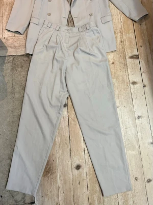 Beige kostym med dubbelknäppt kavaj st. 42 - Snygg beige kostym med dubbelknäppt kavaj och matchande byxor. Kavajen har klassisk krage, tre fickor och knappar framtill. Byxorna har hög midja och raka ben. Perfekt för en stilren look.