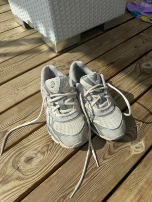Asics GEL NYC Cream - Snygga sneakers från Asics i vitt och ljusgrått med mesh och syntetdetaljer. Skorna har klassisk snörning, vadderad plös och sula med bra grepp. Perfekta för dig som gillar sportig och clean stil.