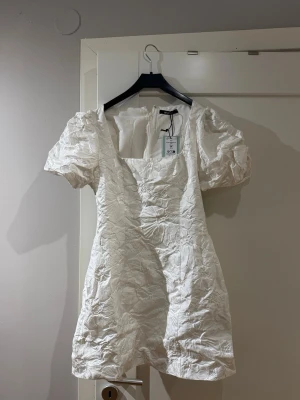 Vit kort klänning med puffärm  - Kort vit klänning med puffärmar och fyrkantig ringning. Klänningen har ett blommigt mönster i tyget och är tillverkad i ett glansigt material. Klänningen är helt ny och oanvänd, med lapp kvar. Sååå himla fin. Kan användas vid finare tillfällen, student, examen etc. Säljes pga fel storlek❤️