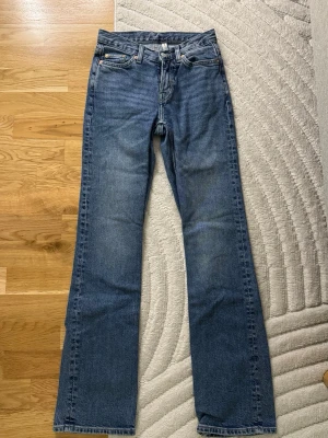 Blå raka jeans med medelhög midja - Säljer ett par klassiska blå jeans med rak passform och medelhög midja. Jeansen har fem fickor, bälteshällor och knappgylf. Materialet är kraftig denim i bomull och färgen är mellanblå med lätt slitning. Perfekta för en avslappnad och stilren look.