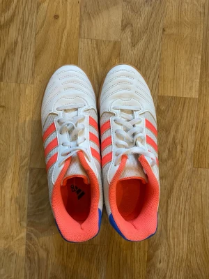 Vitblå futsal inomhusskor Adidas - Snygga futsalskor från Adidas i vitt med röda ränder och blå detaljer vid hälen. Skorna har klassisk snörning, låg profil och innersula i rosa mesh för extra komfort. Yttersulan är i gummi, perfekt för inomhusspel. Storlek 36 2/3
