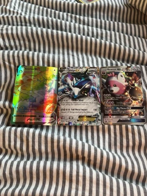 Pokémonkort Snorlax VMAX, Dialga EX, Bewear GX - Tre Pokémonkort: Snorlax VMAX med regnbågsglans, Dialga EX med blå och silver detaljer samt Bewear GX med svart och rosa motiv. Alla korten har holografiska effekter och är perfekta för samlare eller Pokémonfans.