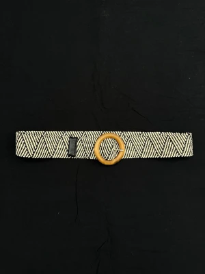 Randigt bälte med träspänne - Bälte/skärp med svart och beige randigt mönster och ett runt spänne i trä. Bältet har en bred design och är stretchigt. 