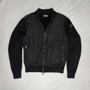 Moncler Cardigan  - Snygg svart Moncler Cardigan med nylon front och stickade ärmar. Koftan har ribbade muddar, hög krage och två dragkedjeförsedda fickor framtill. Perfekt för dig som gillar en sportig men ändå stilren look.