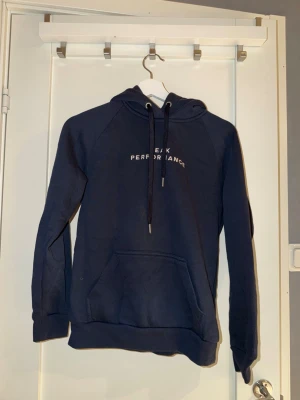 Mörkblå hoodie Peak Performance - Snygg mörkblå hoodie från Peak Performance med broderad logga på bröstet. Justerbara snören i huvan och ribbade muddar vid ärmar och nederkant. Tillverkad i mjukt bomullsmaterial för skön känsla.
