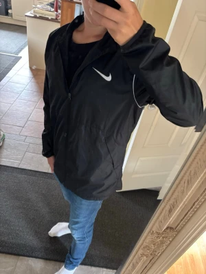 Svart Nike vind/regnjacka med vit logga - Snygg svart vindjacka från Nike med vit logga på bröstet och vita detaljer längs ärmarna. Jackan har dragkedja framtill och en avslappnad passform. Perfekt för blåsiga dagar och enkel att matcha med jeans eller träningskläder.