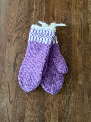 Lila stickade vantar med vitt mönster - Ett par handstickade vantar i lila med vita detaljer upptill. Vantarna har ett klassiskt tumgrepp och ribbad kant med dekorativt mönster. Perfekta för kalla dagar och ger en mysig känsla.