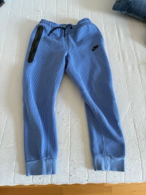Blå Nike mjukisbyxor med dragkedja - Säljer ett par blå mjukisbyxor från Nike med svart logga på vänster ben och en svart dragkedjeförsedd ficka. Byxorna har ribbade muddar vid bensluten och är gjorda i ett strukturerat, rutigt tyg. Perfekta för chill eller träning.