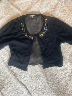 Svart bolero med stenar från M&Co - Kort svart bolero från M&Co Boutique med glittriga stenar och pärlor längs den rundade urringningen. Tunn och mjuk kvalitet, perfekt att slänga över en topp eller klänning. Trekvartsärm och öppen framtill för en chill och elegant vibe.