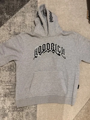 Grå HOODRICH hoodie med tryck - Snygg grå hoodie med svart och vitt HOODRICH-tryck på bröstet och text på huvan. Klassisk känguruficka framtill och ribbade muddar vid ärmar och nederkant. Perfekt för en chill och street-inspirerad look.